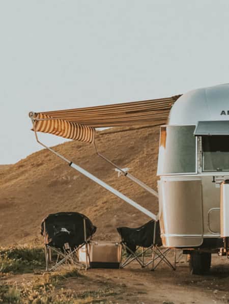 A silver Airstream trailer parked at a campsite next to a hill, with an awning and chairs set up on its side.