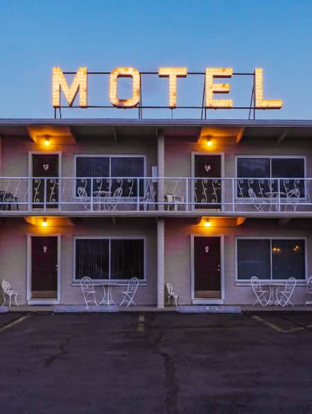 The front view of a motel with a parking lot in front, where each room door is illuminated by a porch light.