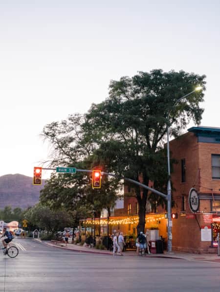 The corner of a busy city street, showing a cross street, pedestrians walking, and a cyclist riding by.