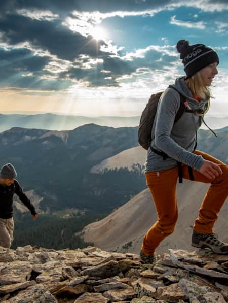 A man and a woman hiking up a rocky mountain trail with mountain ranges visible in the distance behind them.