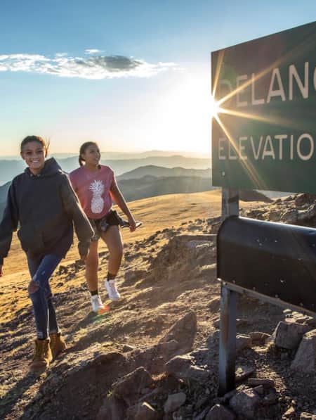Two young girls standing at the summit of a mountain next to a sign that reads 'Delano Peak Elevation 12,169'.