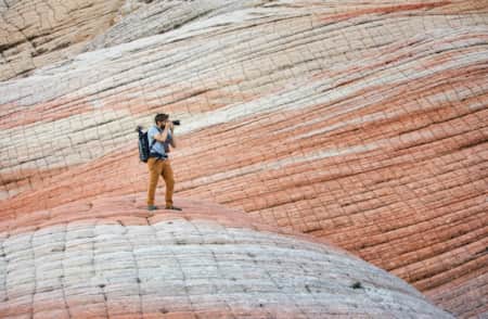 A man with a large camera taking a photo of something in the distance, standing on and surrounded by large rock formations with white and red striations.