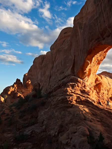 Four hikers standing underneath a natural bridge, illuminated by the sun, amidst a landscape of red rock formations.