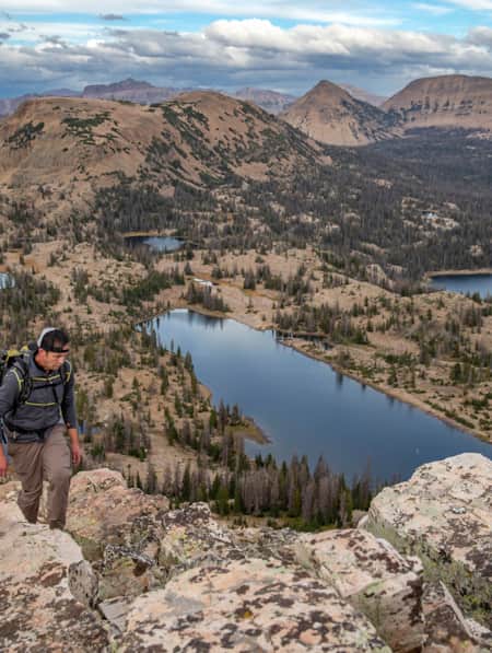A man hiking at the top of a mountain, overlooking multiple lakes scattered across the base and additional mountain ranges in the background.