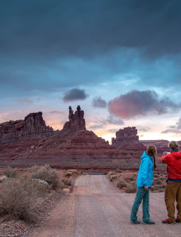 Two people on a gravel road at sunset, with blue hues in the sky, taking a picture of tall, slender rock formations in the distance.