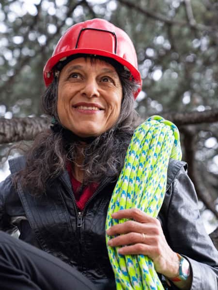 A woman sitting on a tree branch, wearing a red helmet and carrying a neon green rope.