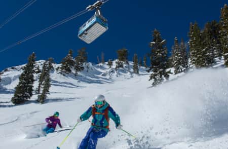 Two skiers descending a mountain with a tram overhead.