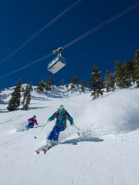 Two skiers descending a mountain with a tram overhead.