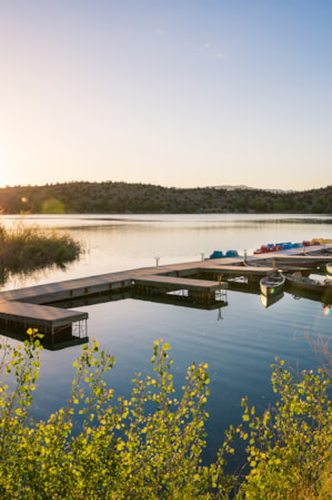A dock on a lake with canoes and pedal boats, and a bright sun setting behind green hills in the background.