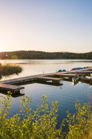 A dock on a lake with canoes and pedal boats, and a bright sun setting behind green hills in the background.