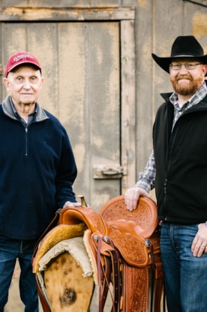 Two men standing on either side of a horse's saddle in front of a barn.