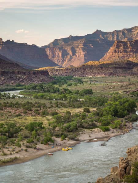 A wide river winding through a rocky canyon of buttes, with some rafters resting at the shore.