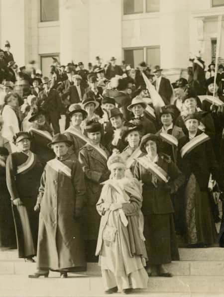 A group of women in long dresses with sashes across their chests, celebrating an event at a government building.