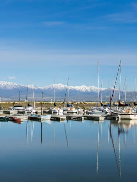 Multiple sailing boats docked on a lake, with snow-capped mountains in the background.