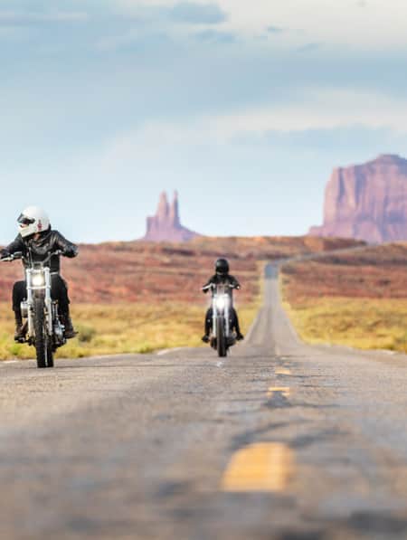 Two people riding motorcycles on a long, straight desert road."