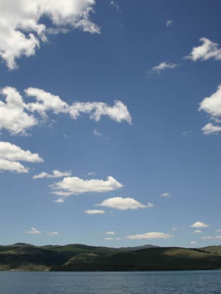 An empty lake with green hills along the shore and a clear blue sky with a few clouds.