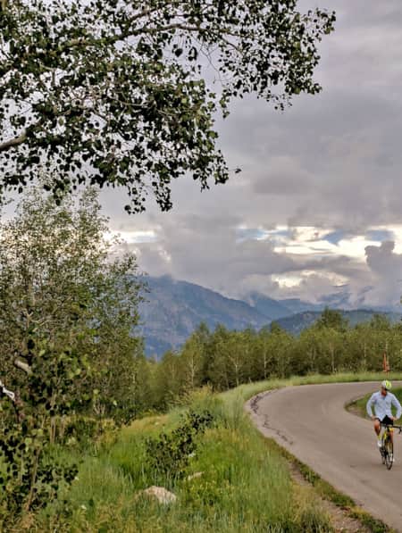 A person cycles on a road surrounded by aspen trees and mountains.