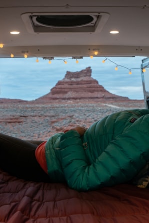 A woman warmly dressed, sitting in a van with a bed set up in the back, gazing out at a large rock formation in the desert.