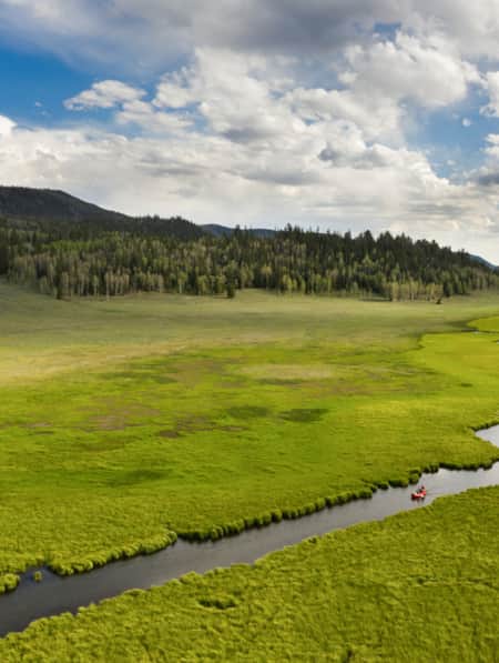 A narrow stream winding through a grassy field, with people kayaking on it and green hills covered with trees on each side.