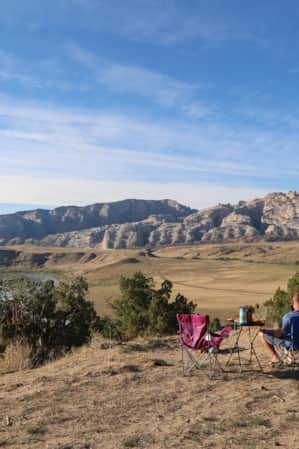 A man sitting in a camp chair with an empty chair beside him and a table in front, gazing out at a river and mountain range.