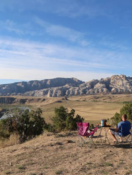 A man sitting in a camp chair with an empty chair beside him and a table in front, gazing out at a river and mountain range.