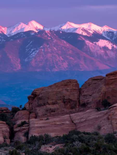 Purple and blue-hued mountains at sunset with snow-capped peaks, and red rock formations prominently featured in the foreground.