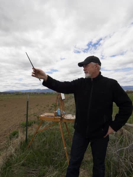 A man in an open field with an easel, holding up his paintbrush.