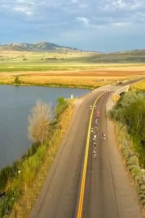 A two-way road paved through a lake, with cyclists racing on it and open fields in the background.