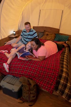 A family lying on a bed, reading a book together inside a canvas tent.