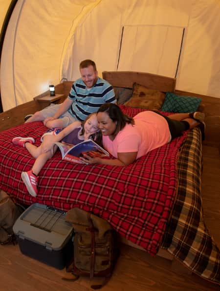 A family lying on a bed, reading a book together inside a canvas tent.