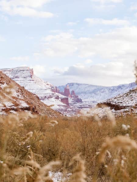A valley filled with hay lightly dusted in snow, nestled at the base of large red rocky mountains with flat plateaus at the top and hoodoos in the background.