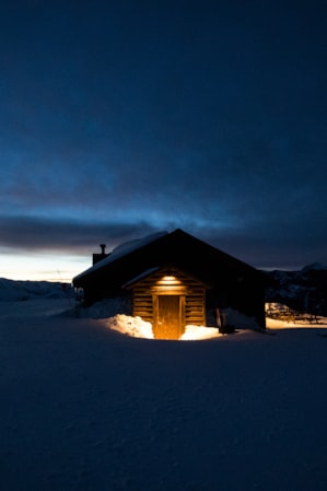 A small lodge in the mountains, surrounded by snow and illuminated by a porch light.