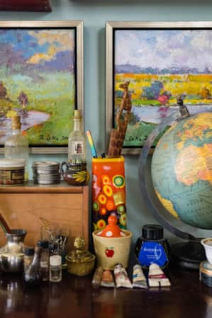 A table with art brushes, water bowls, and other art supplies in front of two paintings depicting nature landscapes.