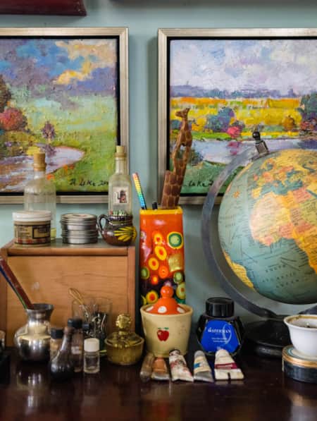 A table with art brushes, water bowls, and other art supplies in front of two paintings depicting nature landscapes.