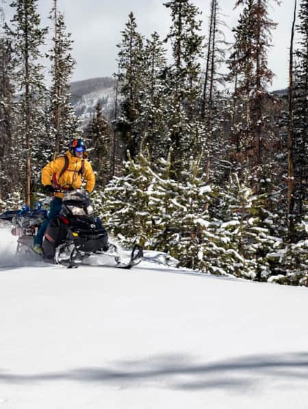 Two people driving snowmobiles on a snowy path through the mountains on a cloudy day.