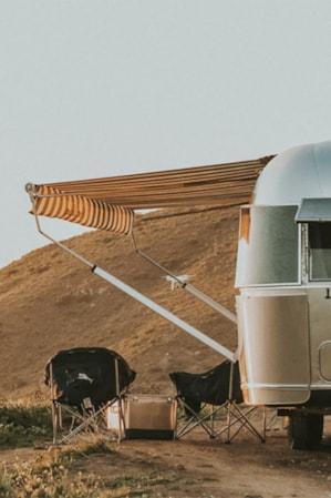 A silver Airstream trailer parked at a campsite next to a hill, with an awning and chairs set up on its side.