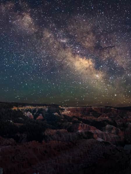 An expansive view of layered rock mountains with trees growing on them, under a starry night sky.