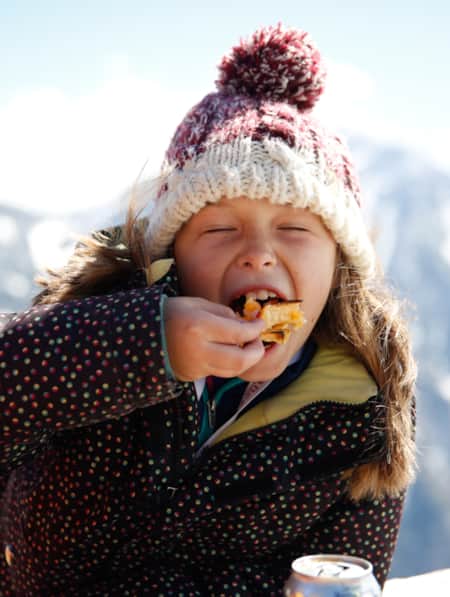 A young girl sitting outside in a ski jacket and hat, taking a big bite of her food at a mountain restaurant.