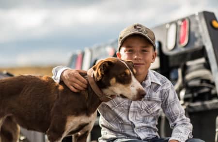 A young boy with his arm around a dog standing beside him.