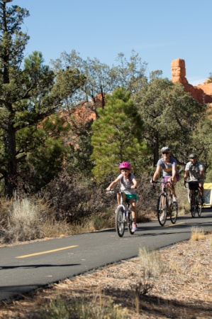 A family of four biking on a paved trail with red rock formations in the distance and trees lining both sides of the path.