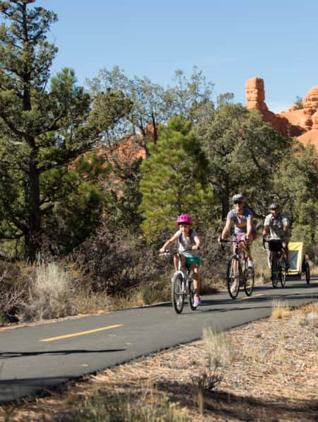 A family of four biking on a paved trail with red rock formations in the distance and trees lining both sides of the path.