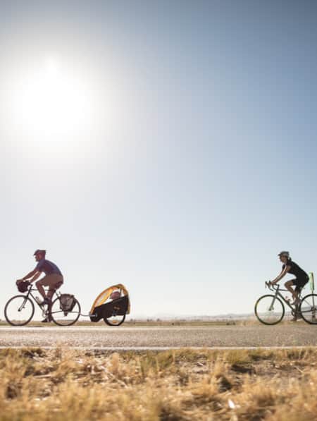 A man on a bicycle towing a bike trailer carrying a child, with a woman riding a bike behind them.