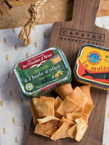 A wooden serving board with two tins of canned fish and crispy pita chips on top.