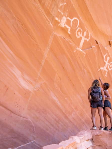 Two women looking up at petroglyphs on a large, flat rock wall.