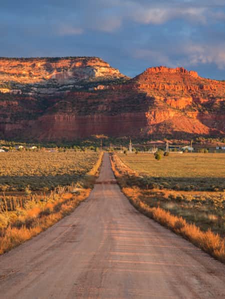 A straight road in the desert leading to a large red rock monument.