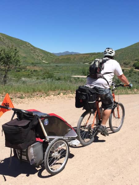 A person biking on a gravel path through green hills, towing a bike trailer.