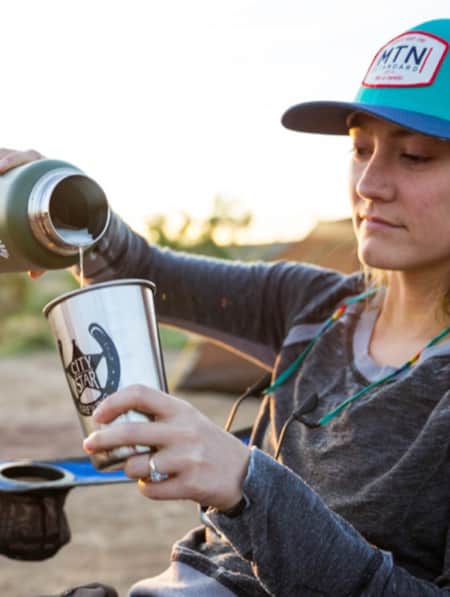 A woman sitting outside in a camp chair, pouring water from a hydro flask into an aluminum drinking cup.