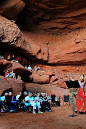 A woman playing the violin by a piano in front of a seated crowd inside a large red rock cove.