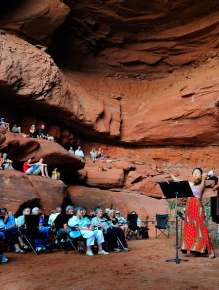 A woman playing the violin by a piano in front of a seated crowd inside a large red rock cove.