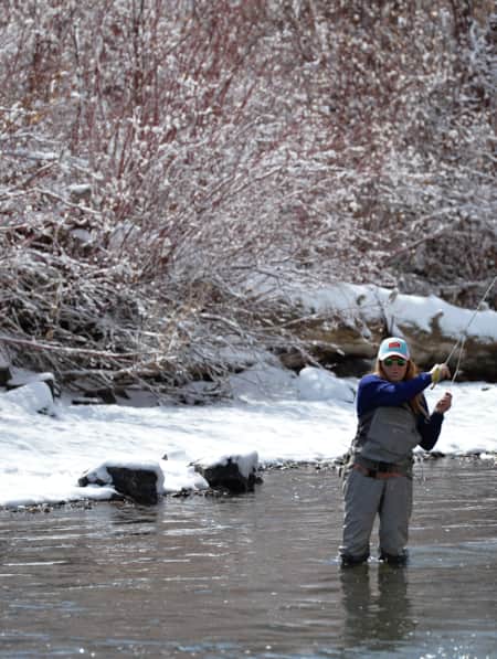 A woman in fly fishing gear standing in a river, with a snow-covered ground along the riverbank